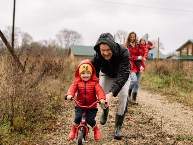 A happy family enjoys a bonding moment outdoors with a child learning to ride a bicycle on a fall day.